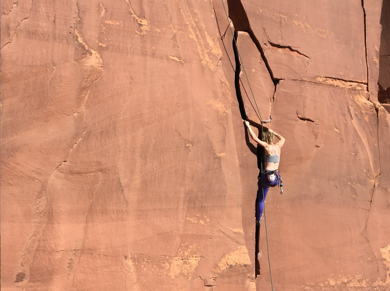 Jessica climbing a crack in Indian Creek on a large slab of red rock. She is wearing a light sports bra and dark leggings with a chalk bag strung around her waist.