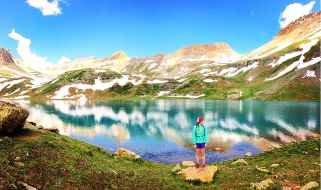Syd Zester in a teal coat and black shorts in front of an alpine lake. The lake features the reflection of the blue sky and mountains behind her.