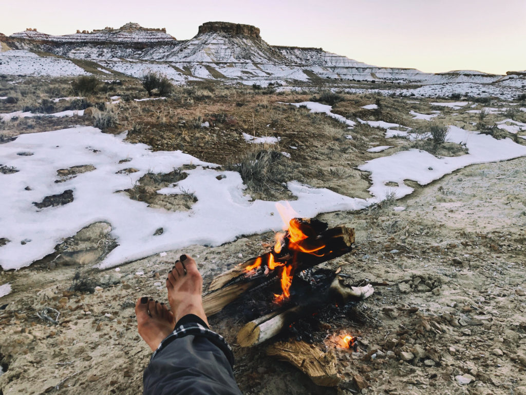 Author Dylan's feet are in the foreground, covered in dirty lines from Chaco sandals. She is seated next to a bonfire with a wide view of the snowy desert.
