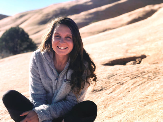 Author Dylan Manderlink sits cross legged and leans toward the camera. She is on her solo journey with mountains in the background. She wears a grey fleece and black pants. Her brown hair is down and reaches past her shoulders.