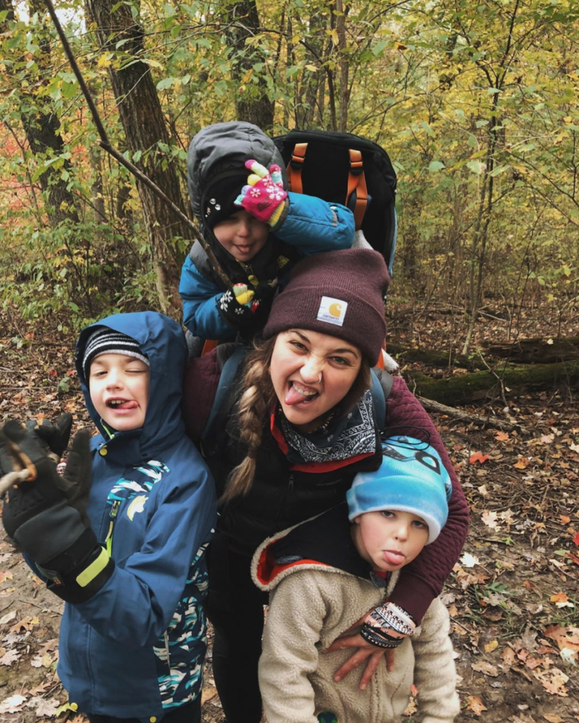 Author Alyx wears her daughter in a child carrier and is flanked by her young sons. They are all sticking their tongues out and having fun.