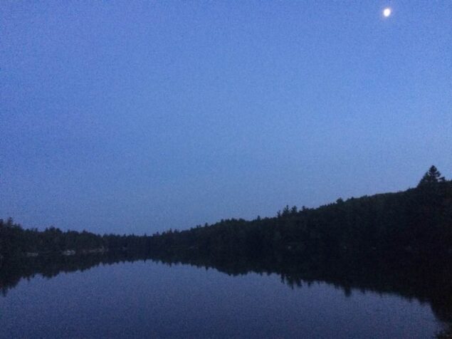 A lake after dusk. There is a small moon in the top right corner, blue sky, black trees, and a dark blue lake.